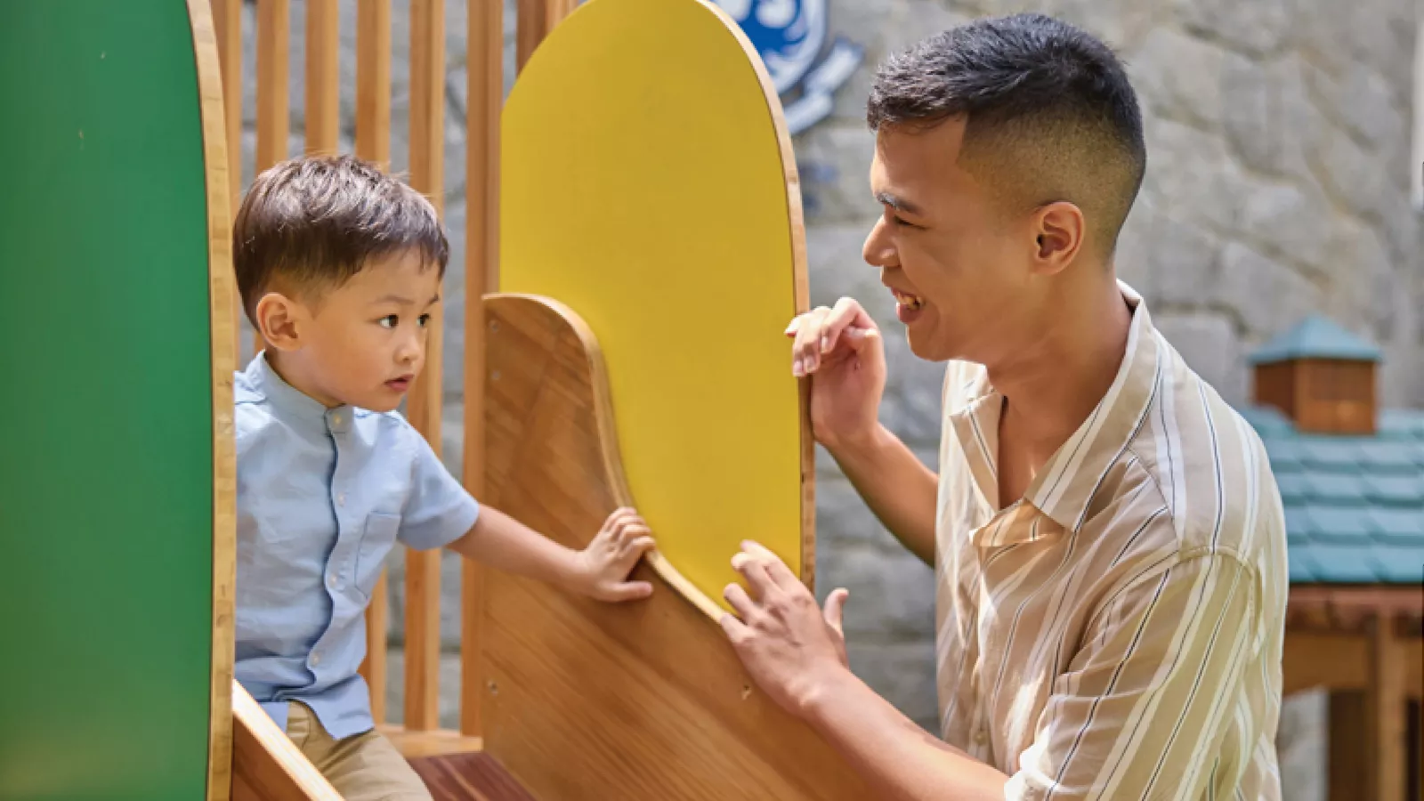A man and child play on a slide, with the man supporting and encouraging the child to go down the slide by himself.