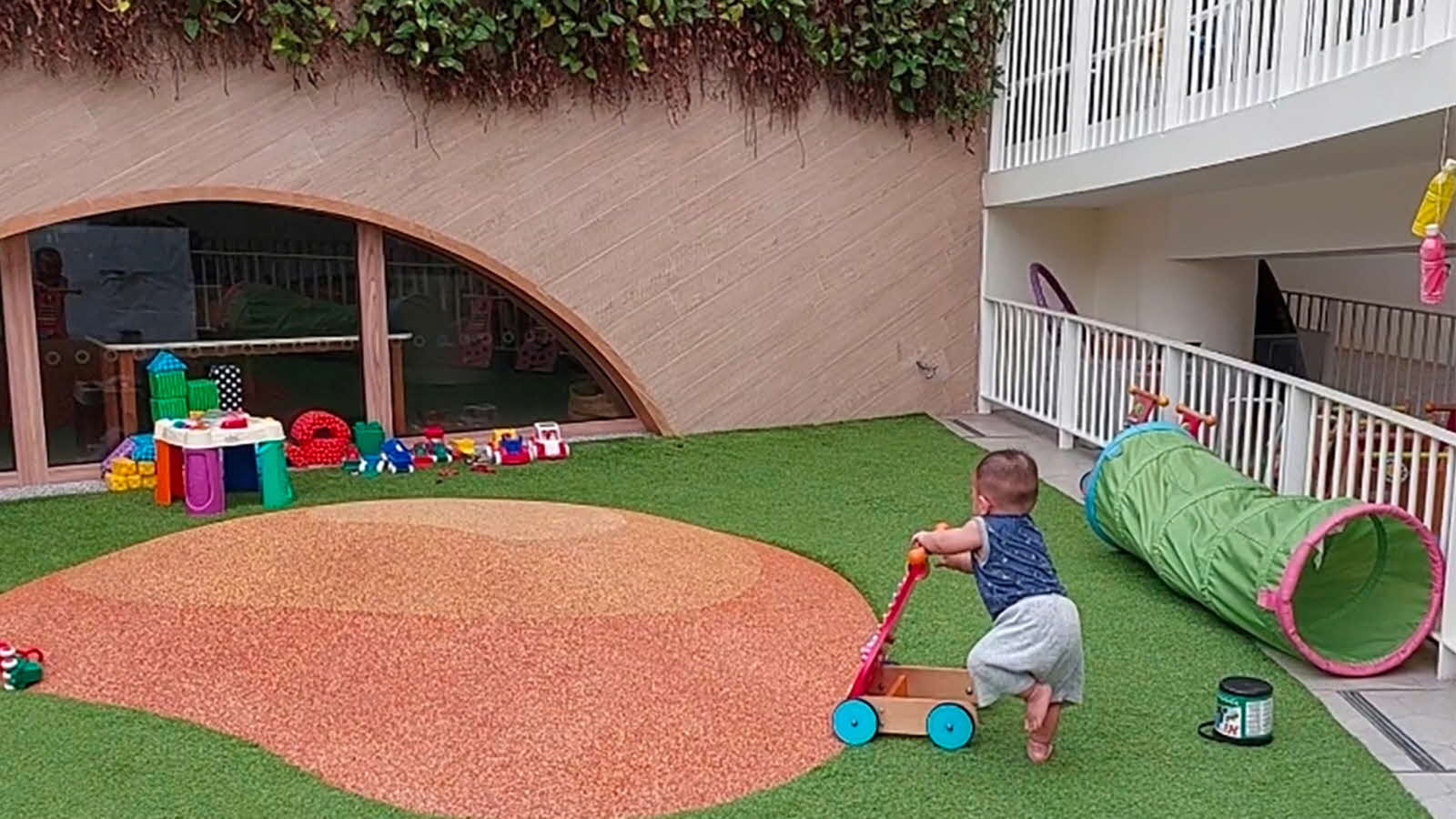 A young child explores an outdoor play area, pushing a cart and taking safe risks while surrounded by various play items.