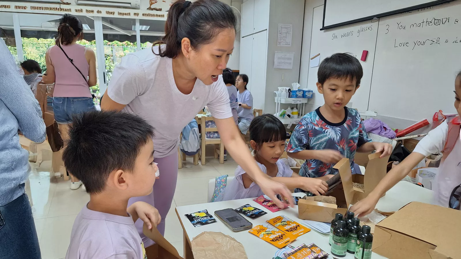 An adult helps children with an SSDB project in the classroom, sorting items on a desk and putting them into care packs.