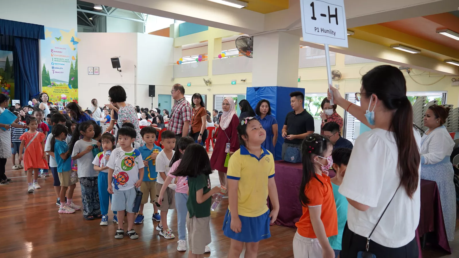 A group of children stand in line at a primary school’s P1 orientation event, with their parents and teachers also present.