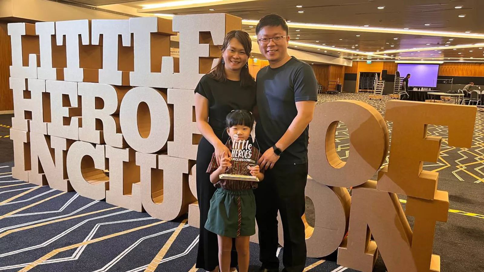 Preschooler Hannah Chung and her parents pose with her award, in front of a sign reading ‘Little Heroes of Inclusion’.