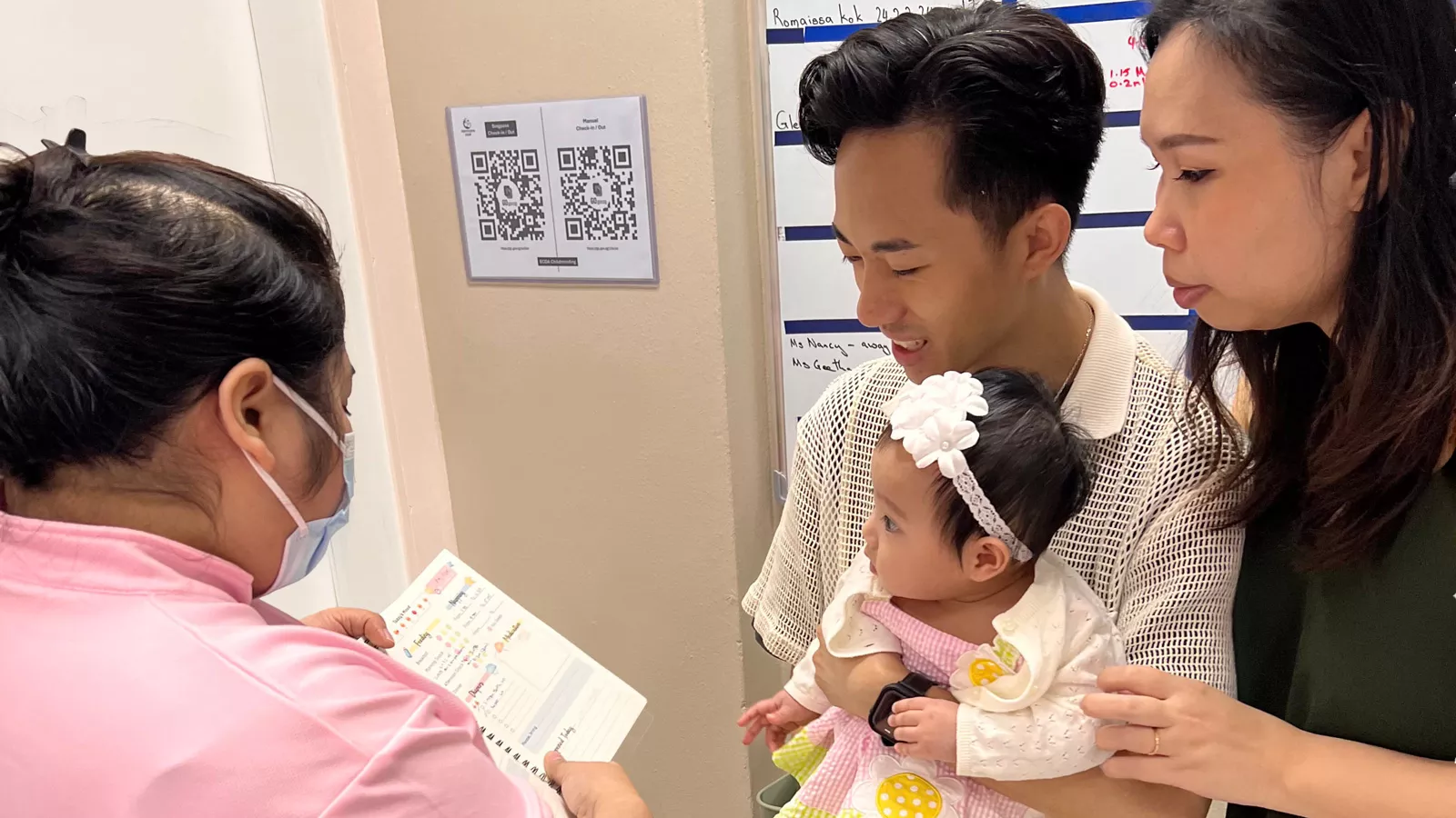 Parents Mr Nathaniel Liu and Ms Norma hold their baby Tabitha while getting updates from a childminder about Tabitha’s day.