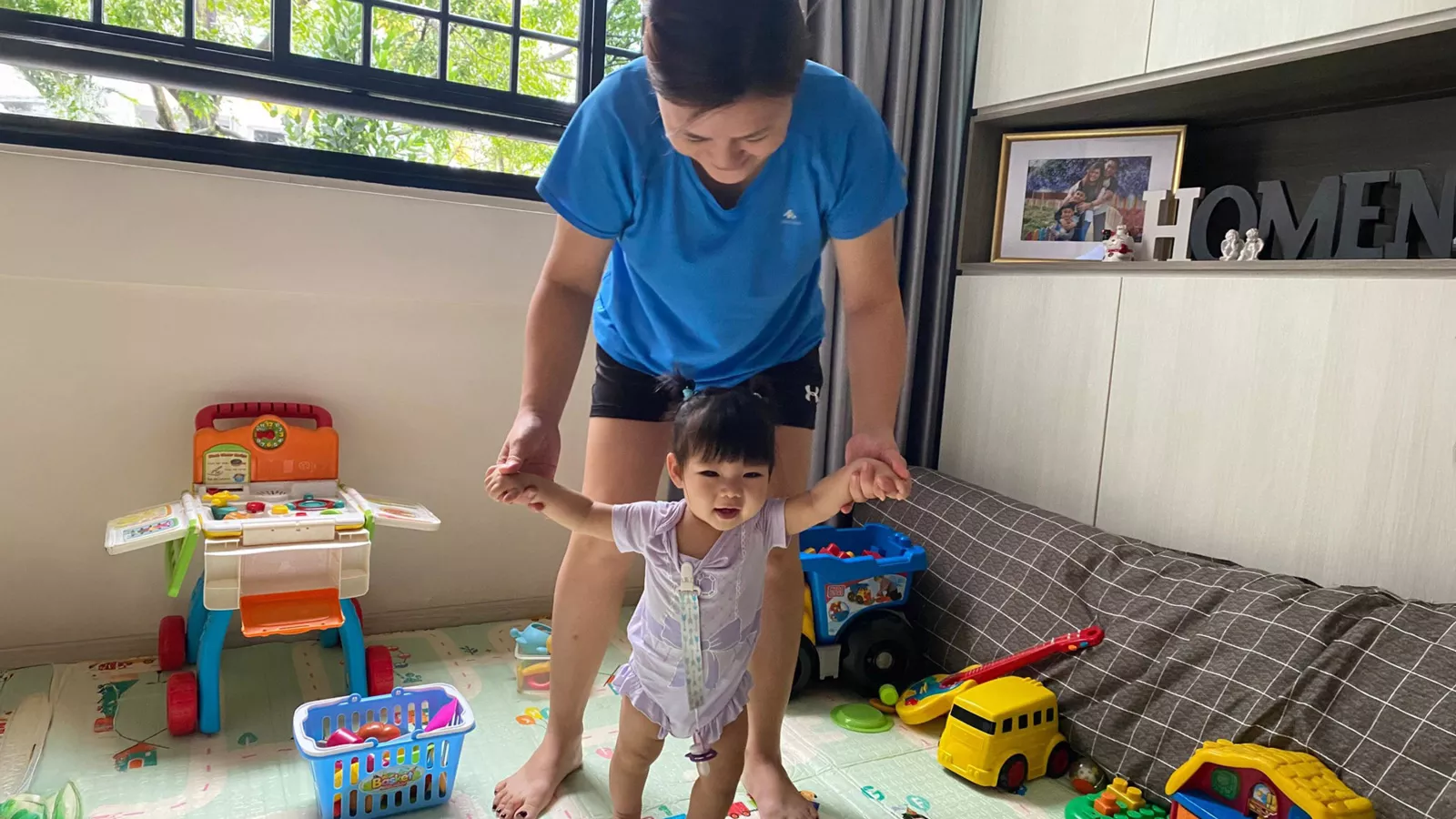 A home-based childminder guides an infant to walk in a cheerful play area filled with toys and cosy furnishings.