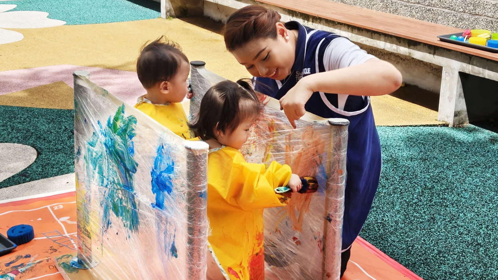 An educator guides infants in an outdoor painting session, fostering creativity, autonomy and self-expression.