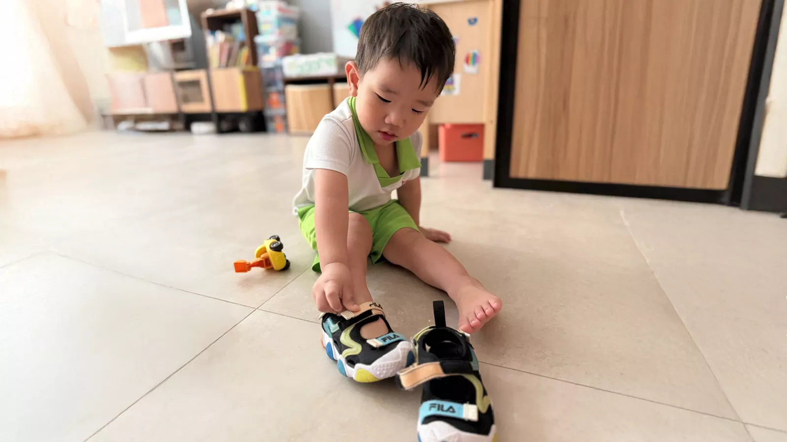 A young boy sits on the floor and puts on his own shoes, demonstrating self-help skills and independence.