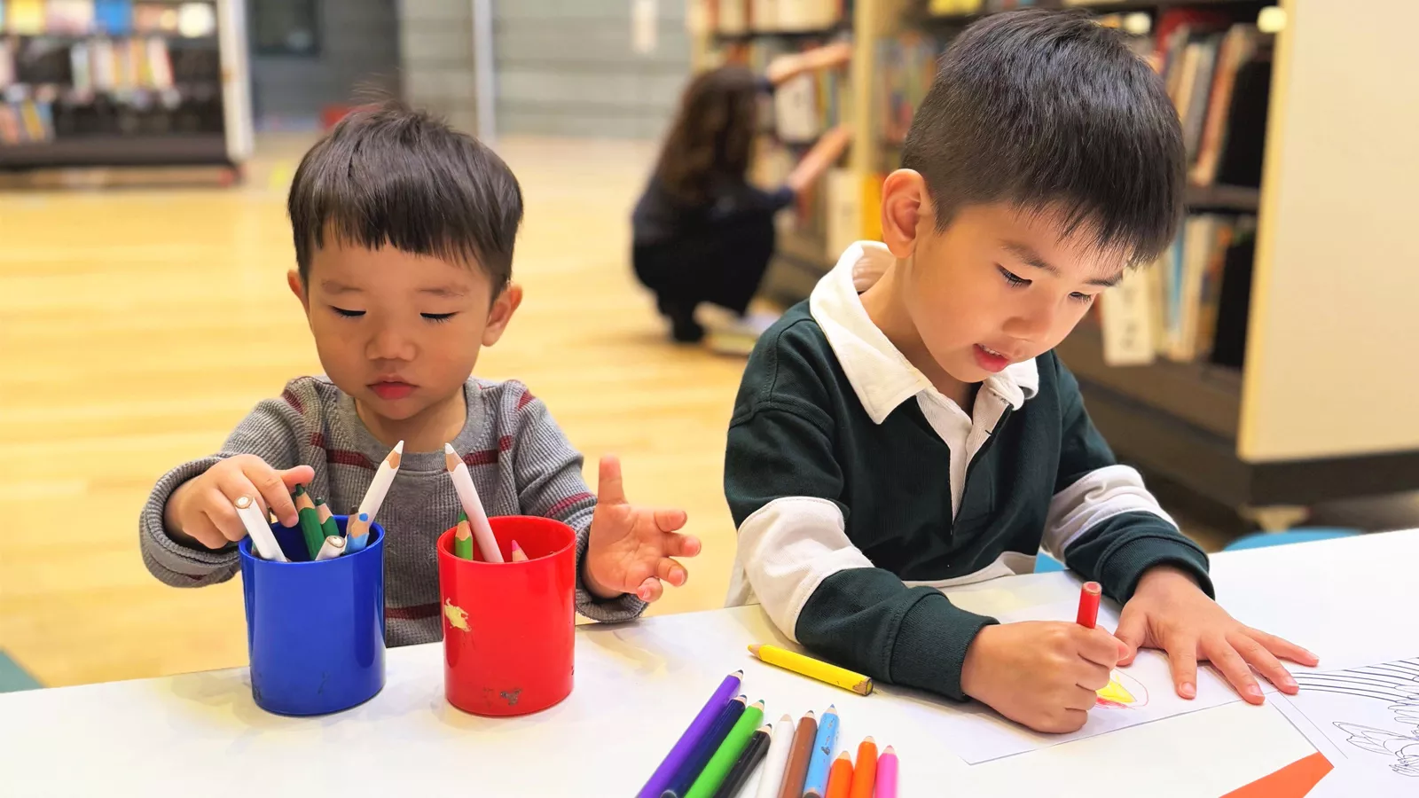 Two boys are focused on drawing at a table in a library, implying autonomy in choosing what activity they want to do.
