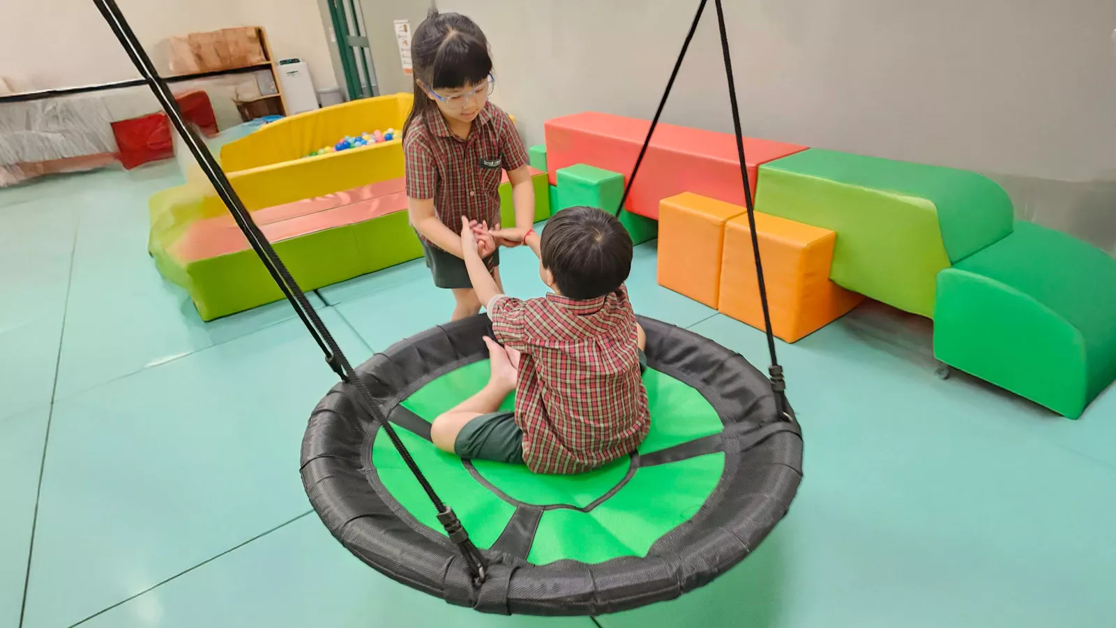 Two children play together in a preschool play gym, with one child kindly helping the other stand up from a swing.
