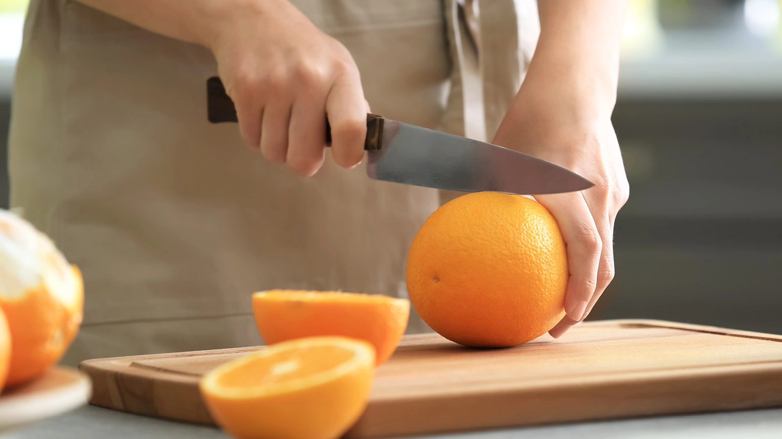 An adult uses a knife to cut an orange on a wooden cutting board.