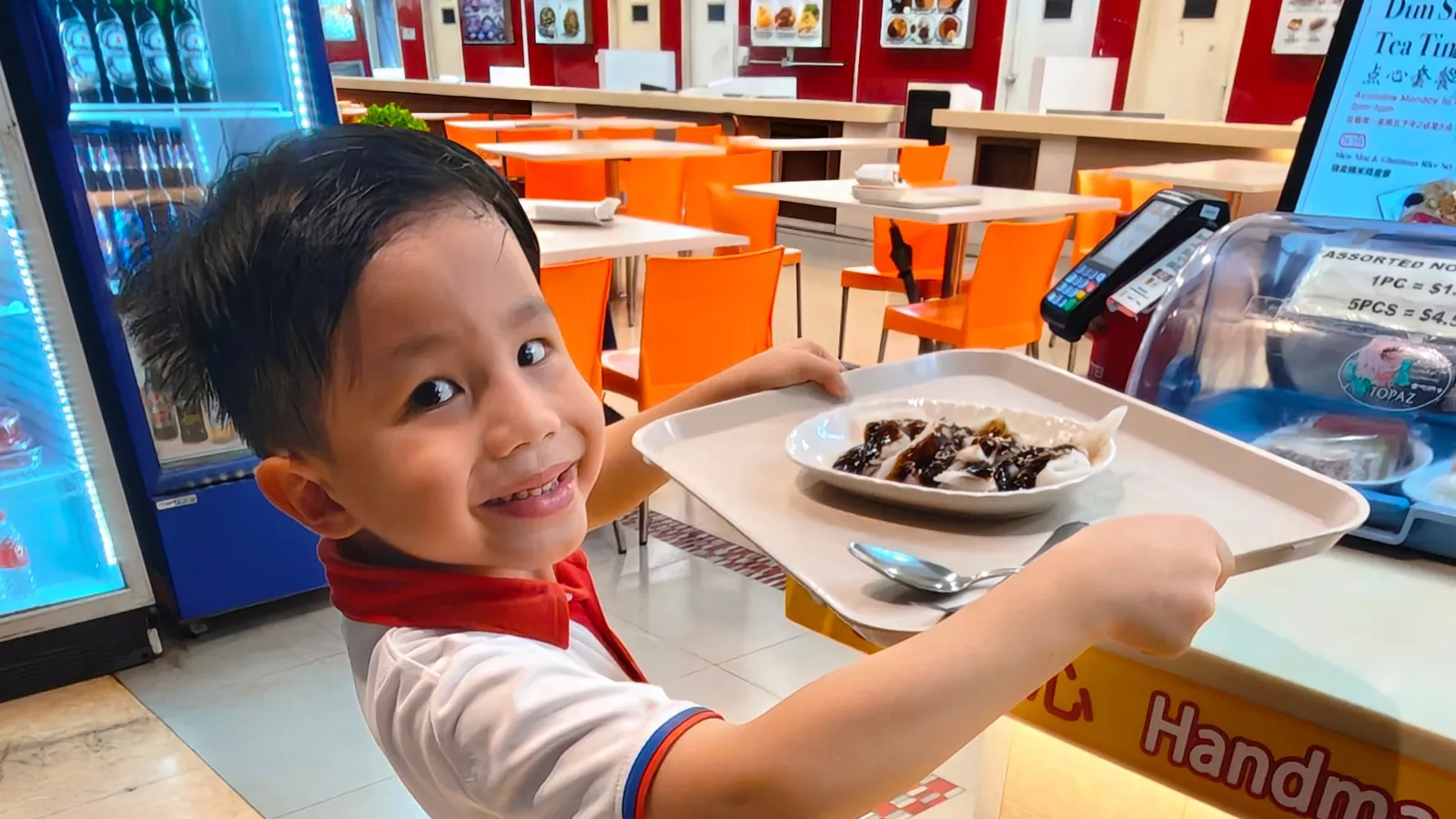 A young boy proudly carries a tray of food in a food court setting, demonstrating independence and self-help skills.